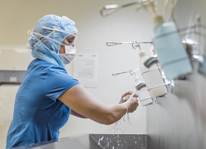 a woman in scrubs and PPE washes her hands at a metal sink